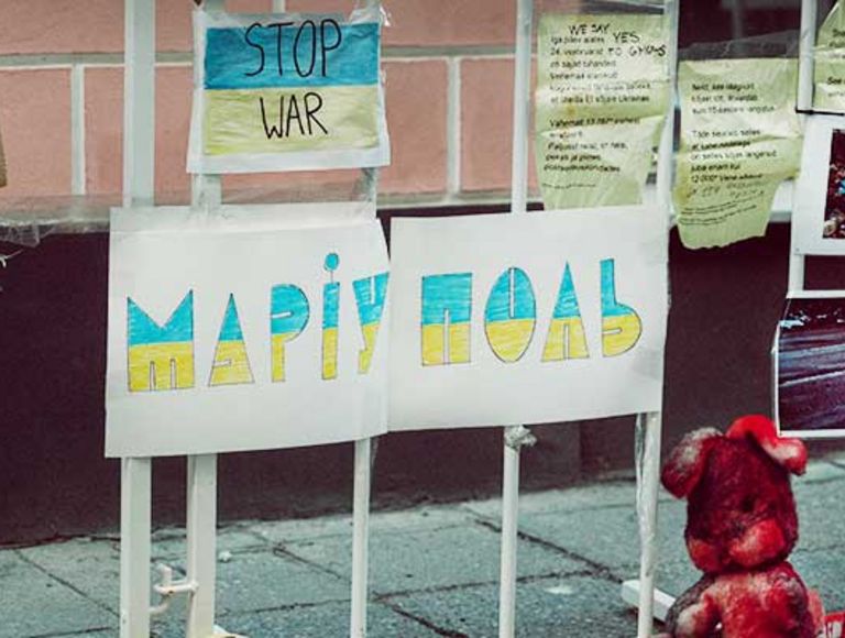 Zaun mit Fotos und Plakaten, darunter der kyrillische Schriftzug "Mariuopol" und eine ukrainische Flagge.
